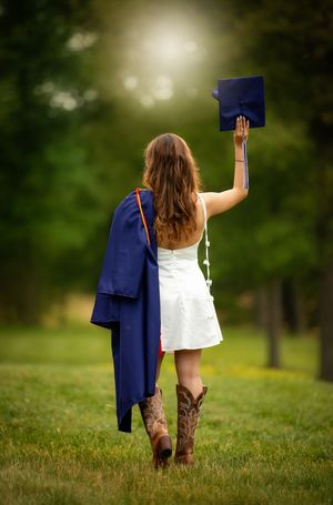 Graduate in white dress and cowboy boots holds cap aloft in lush green field, gown draped over shoulder.