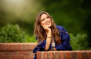 Smiling young woman in blue graduation gown leaning on brick wall with green bokeh background.