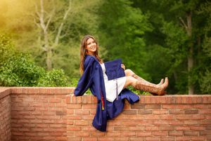 Smiling graduate in navy cap and gown sitting on brick wall with green trees in background.