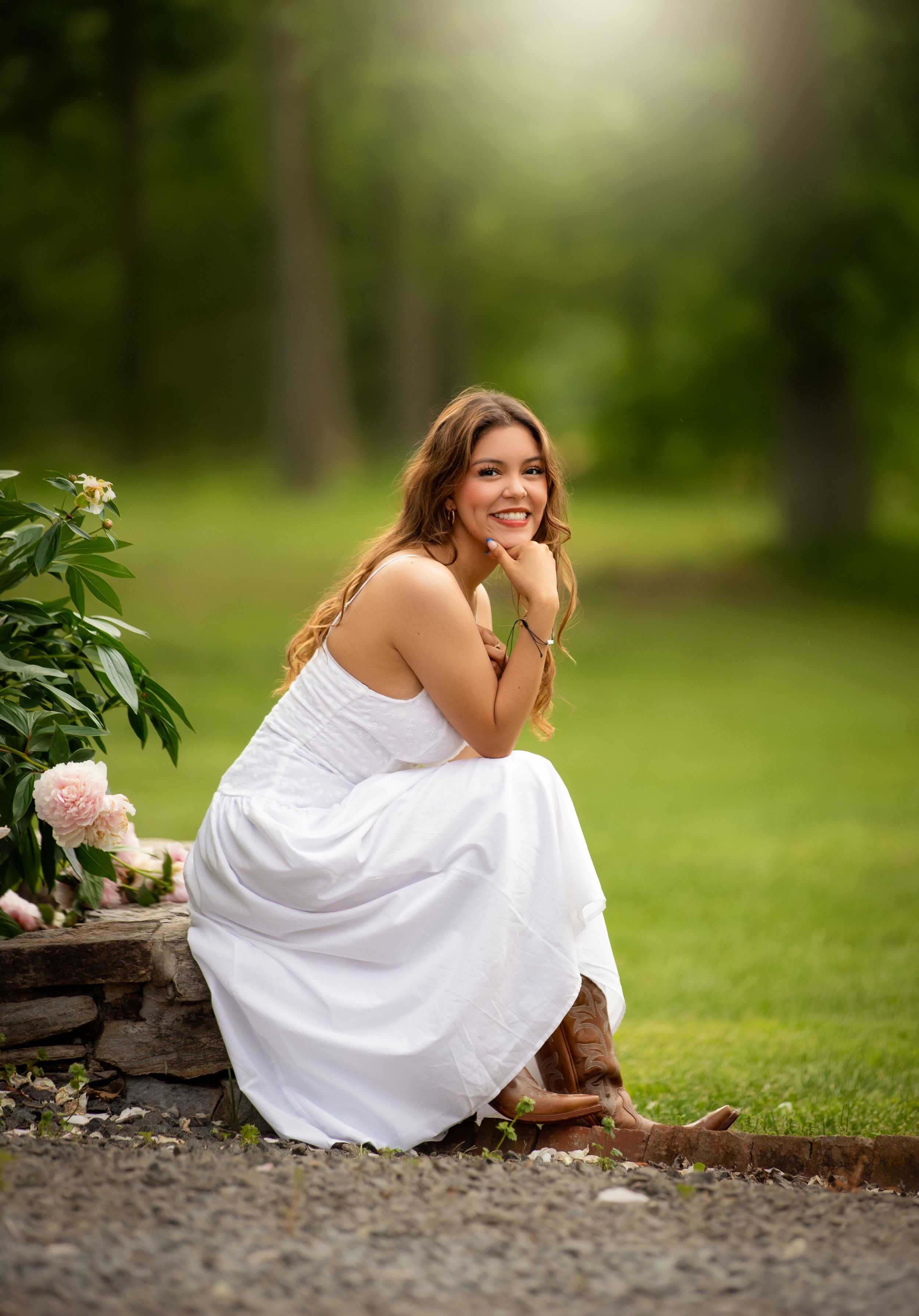 Young woman in white dress and cowboy boots sits gracefully outdoors, smiling beside pink flowers in a lush green setting.