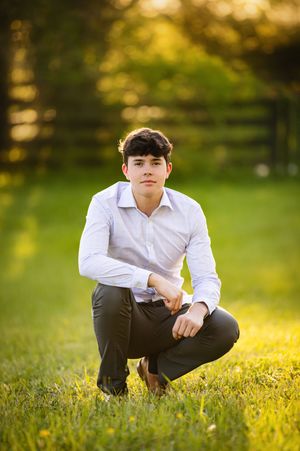 Student in white shirt sits thoughtfully in sunlit grass field during golden hour photography session.