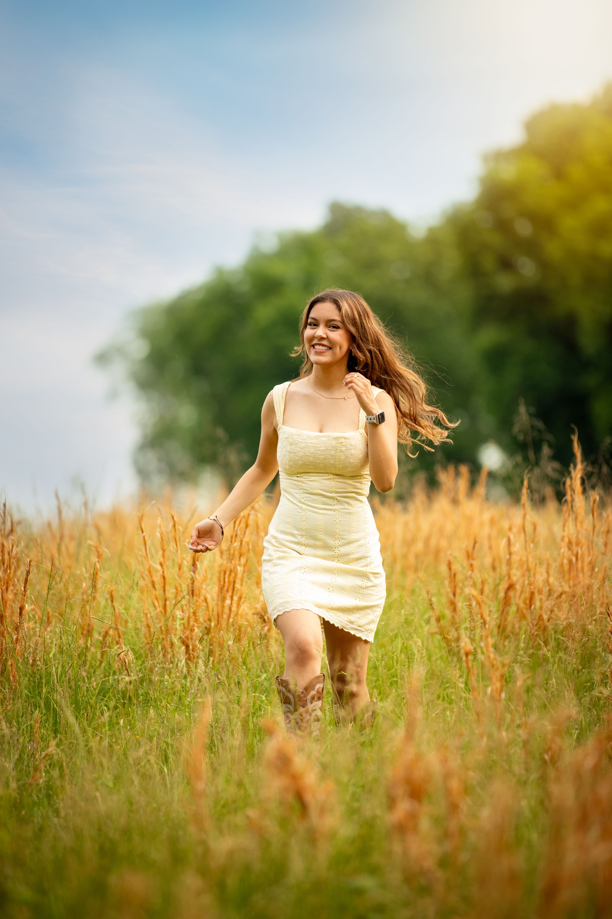 Young woman in yellow dress and cowboy boots running through golden field with trees and blue sky.