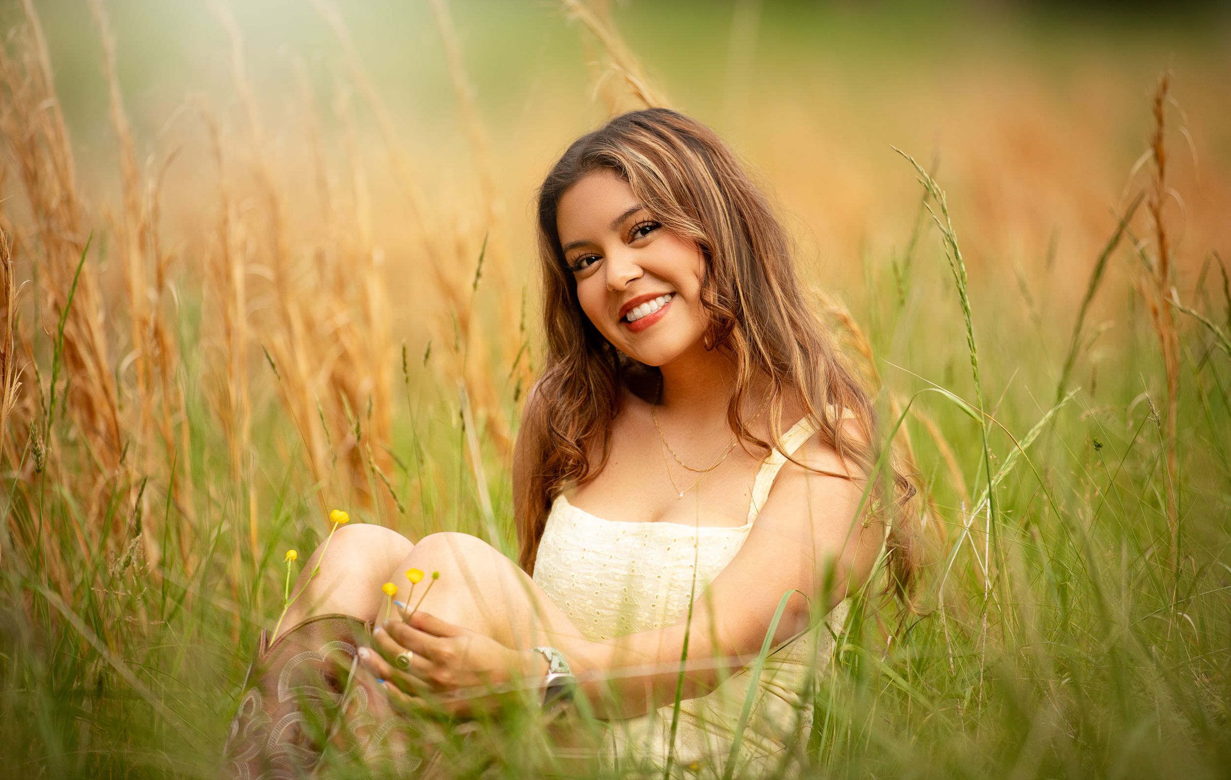 Smiling young woman with long brown hair sitting in tall golden grass field wearing a white dress on a sunny day.