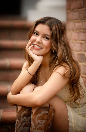 Smiling young woman with long brown hair wearing cowboy boots and a light dress, sitting on brick steps.
