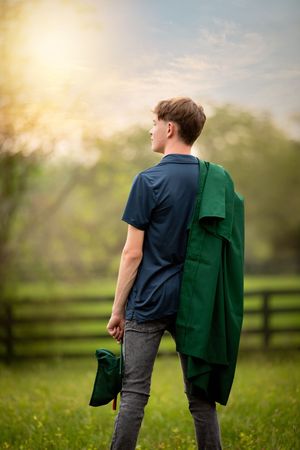 Person wearing navy t-shirt and jeans stands in grassy field carrying green garment in golden sunset light.