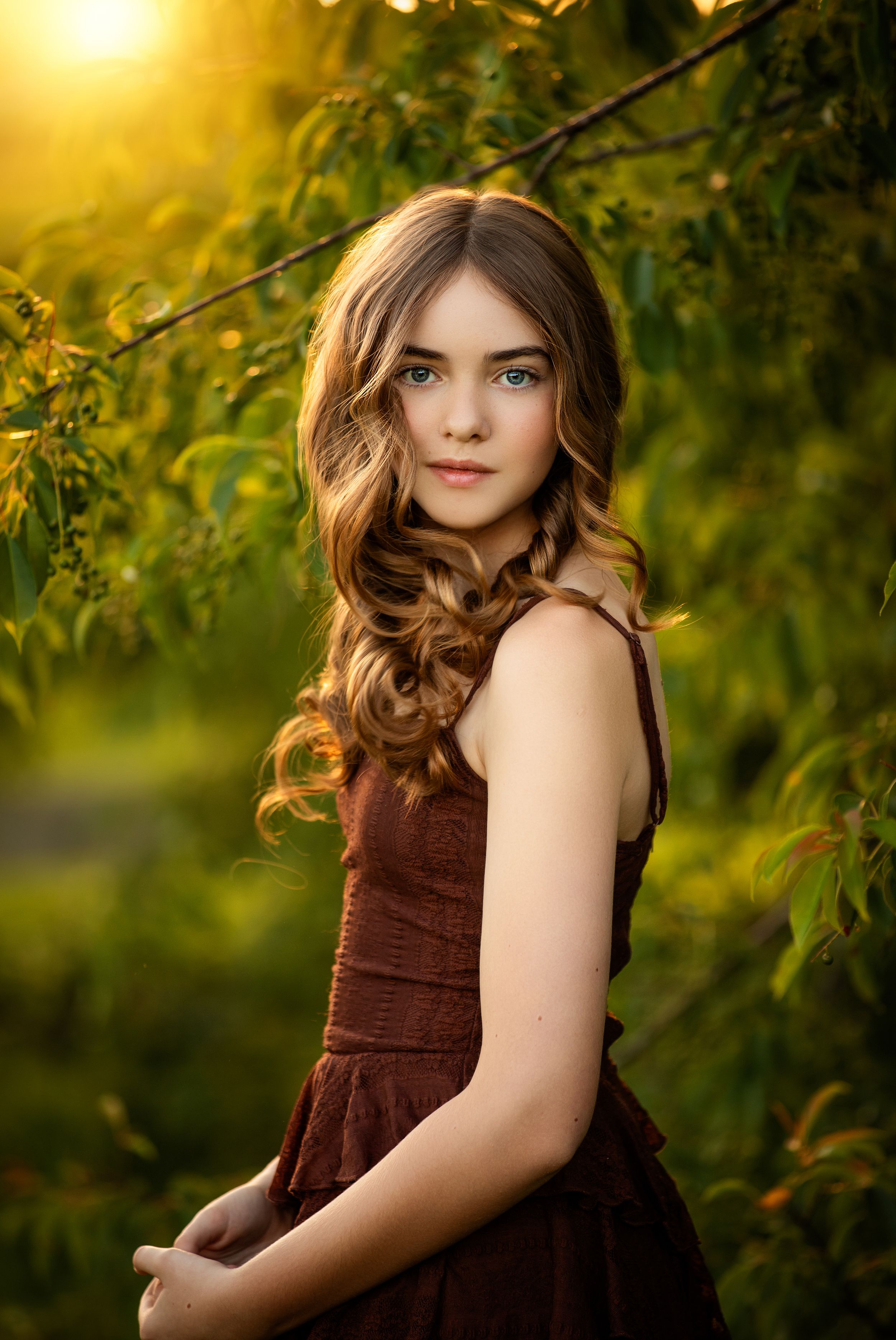 Young girl with curly hair in a burgundy dress poses outdoors in golden sunset light among green leaves.