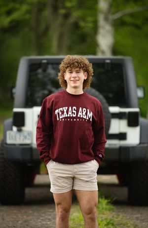 Person wearing maroon Texas A&M sweatshirt and khaki shorts stands outdoors in front of parked vehicles.