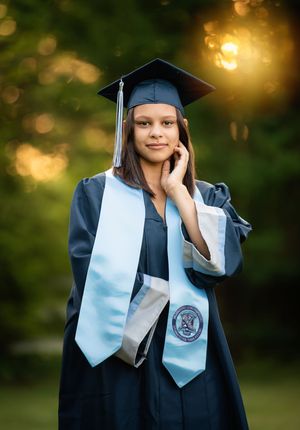 Graduate in cap and gown poses outdoors with glowing bokeh background during sunset photo session.