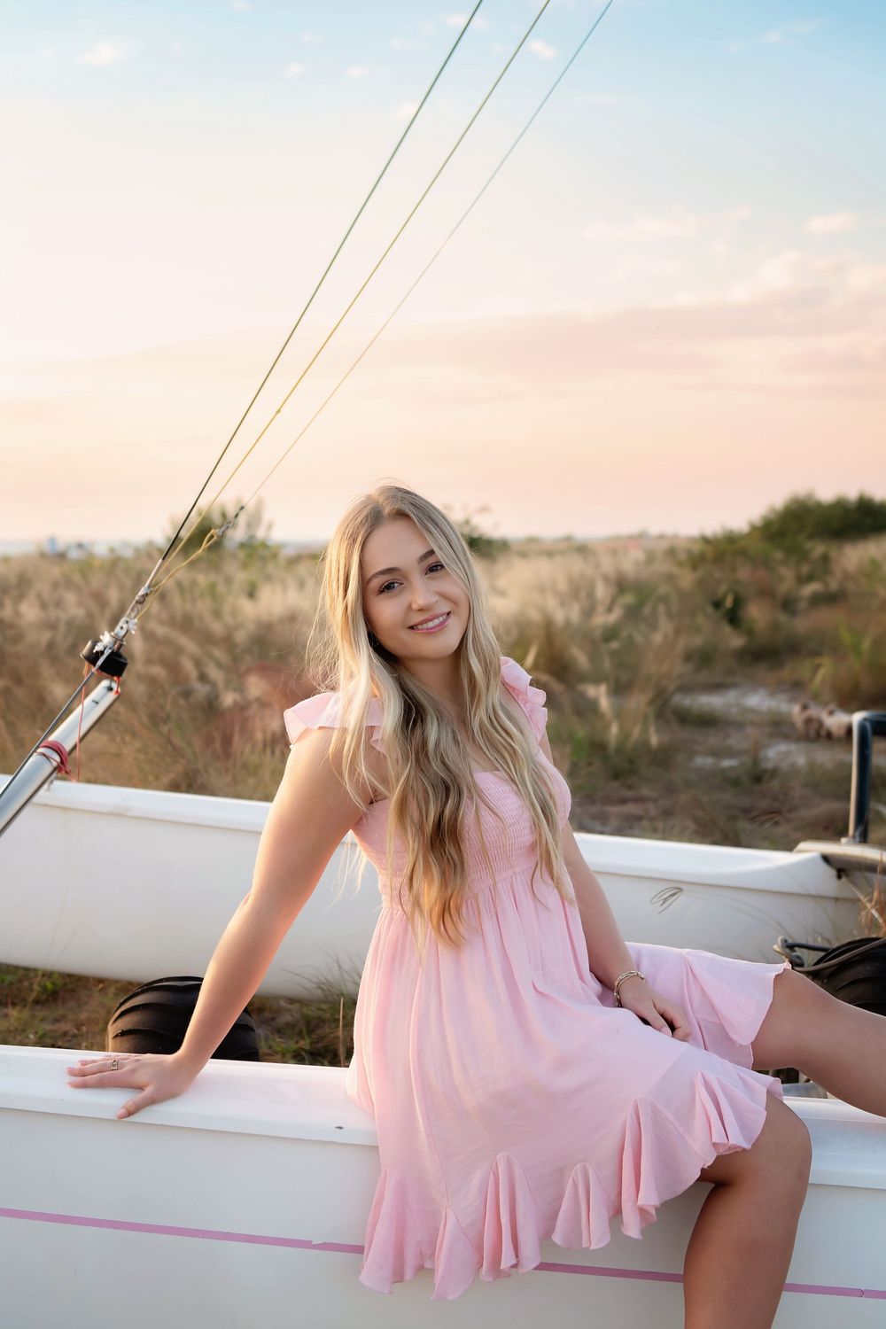 A woman in a pink sundress sits on a sailboat during sunset with coastal scenery in the background.