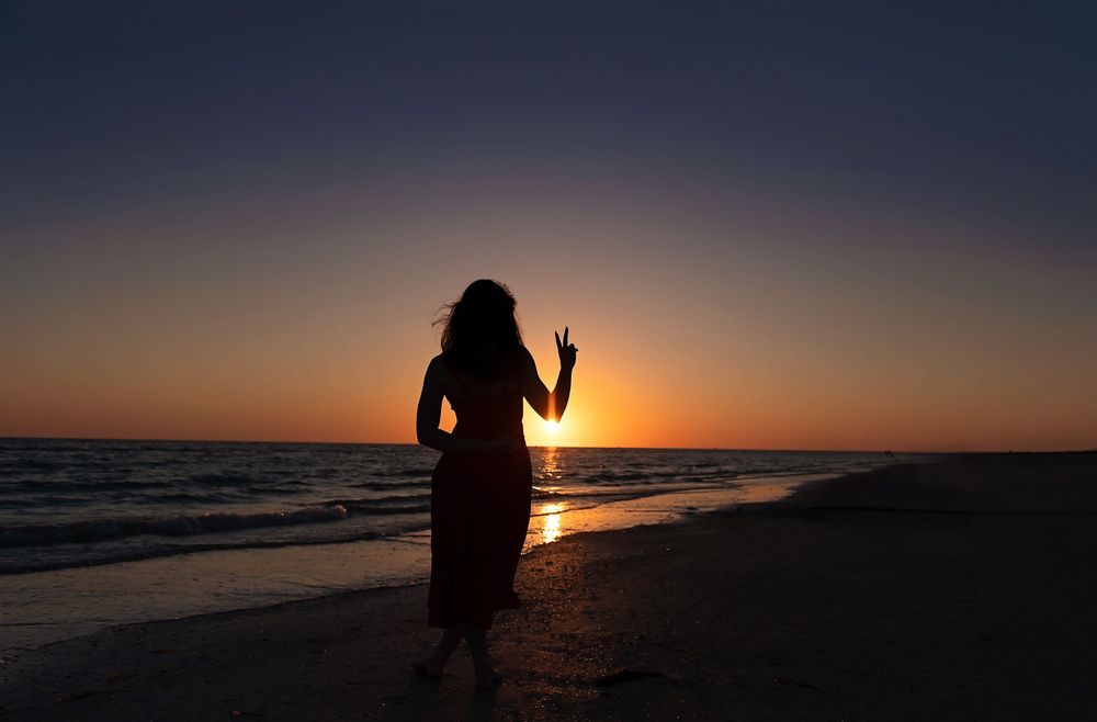 Silhouetted figure stands on beach at sunset with waves crashing along shoreline in dramatic golden light.