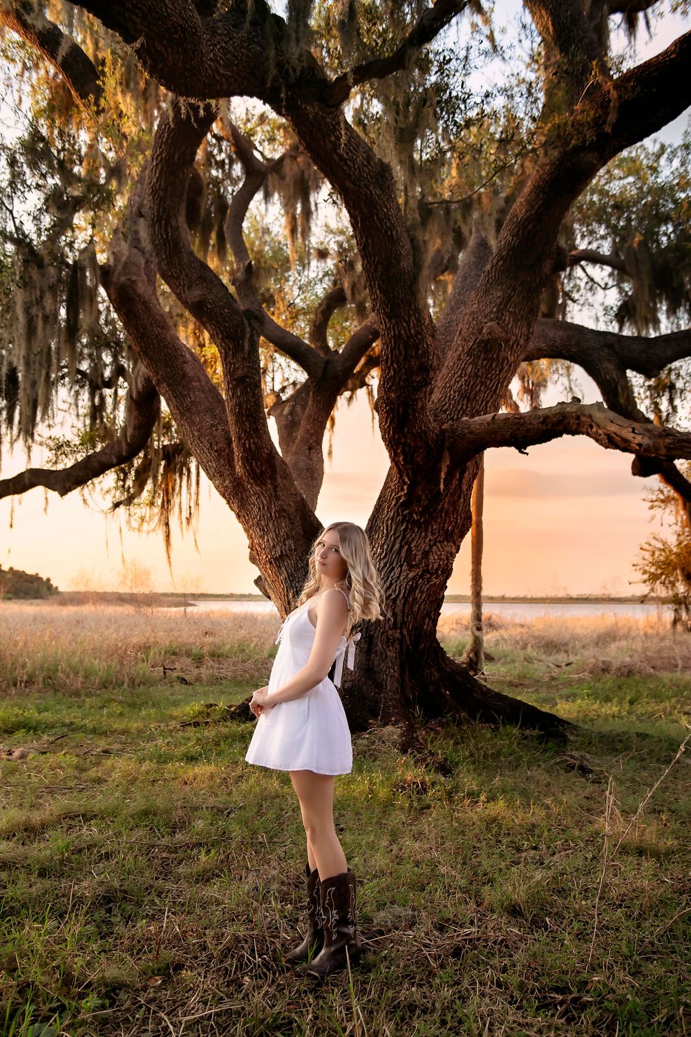 Young woman in white dress and cowboy boots stands beneath a moss-draped oak tree at golden sunset.