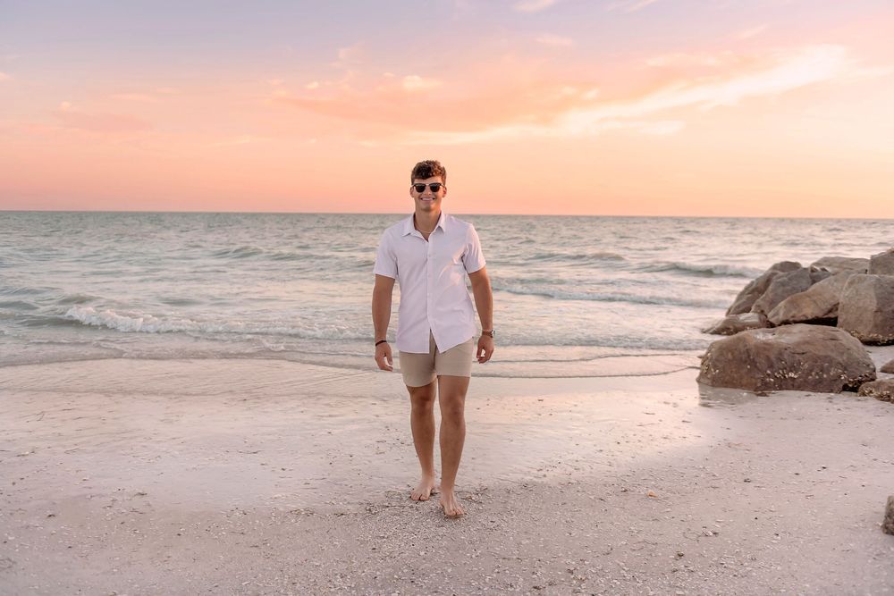 Man in white shirt and shorts walking barefoot on a sandy beach at sunset with pink and orange skies.