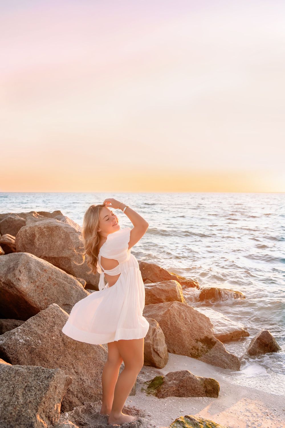 A person in a white dress poses gracefully on coastal rocks during sunset by the ocean.