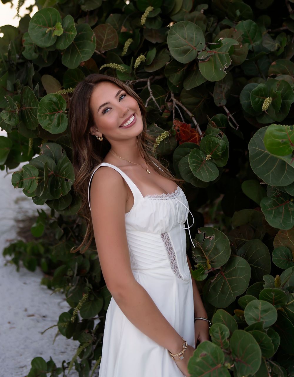 Smiling brunette woman in white sundress posing outdoors against lush green tropical foliage.