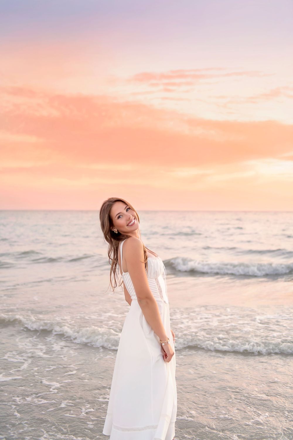 Young woman in white dress smiling at beach during pink sunset, waves behind her.