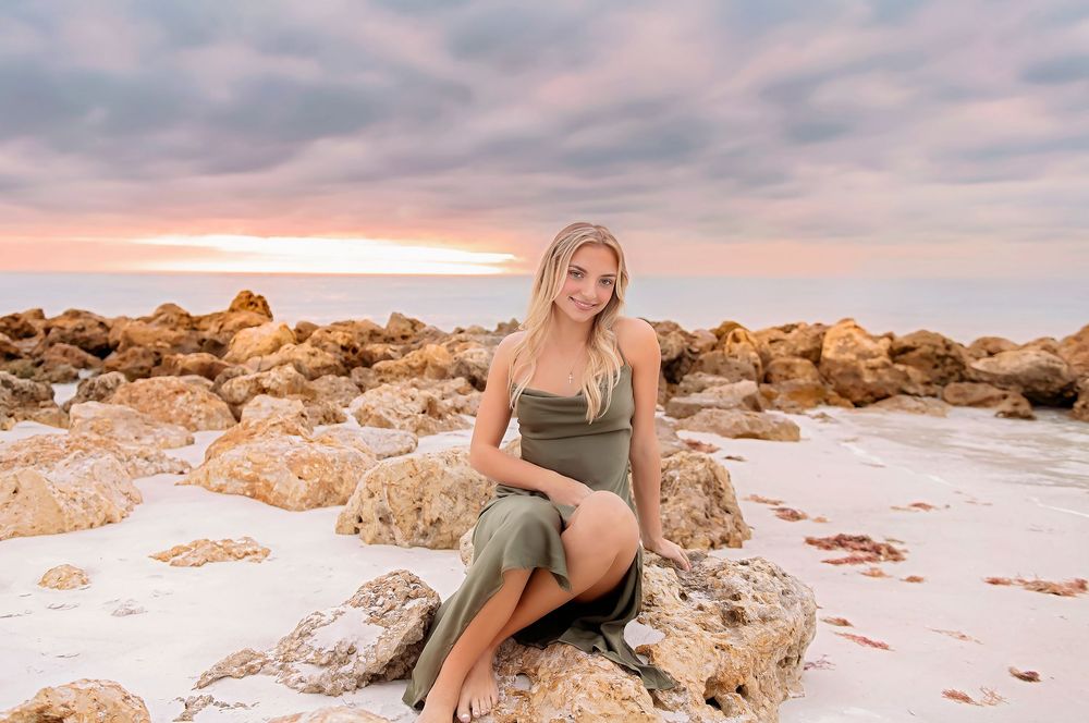 A person poses on rocky coastal cliffs during a vibrant pink and orange sunset by the ocean.