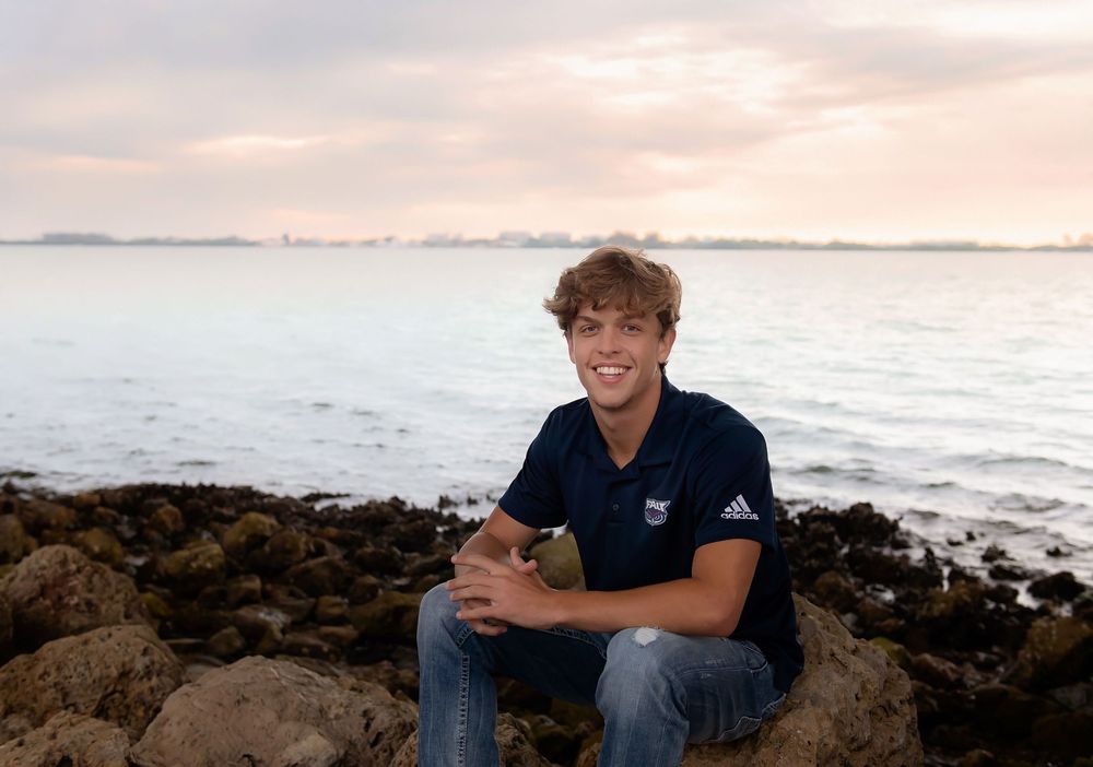 A person in casual clothing sits on rocky coastal terrain at sunset overlooking the ocean waves.