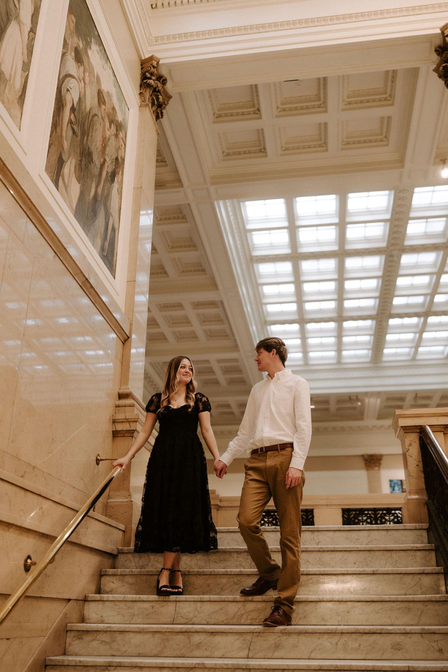 Couple walking down marble staircase holding hands beneath skylight in grand historic building.