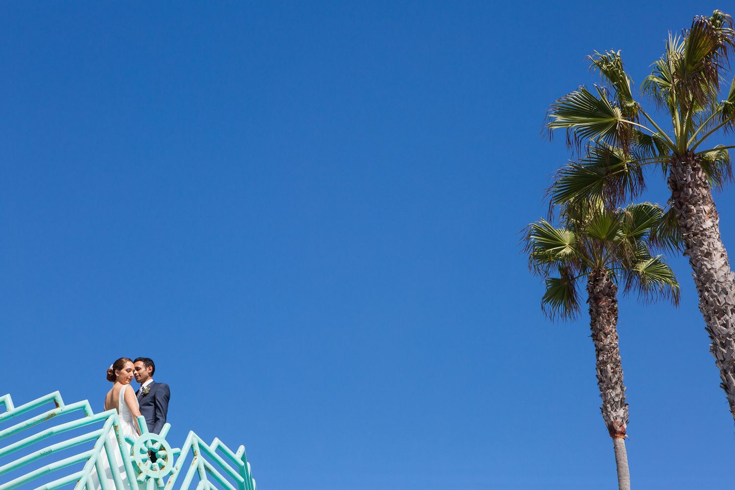 Artistic and unique wedding photo of a bride & groom high up on a balcony surrounded by a clear, blue sky with palm trees opposite them