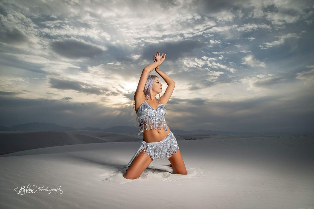 A dancer in a sparkly costume poses gracefully on white sand dunes under a dramatic cloudy sunset sky.