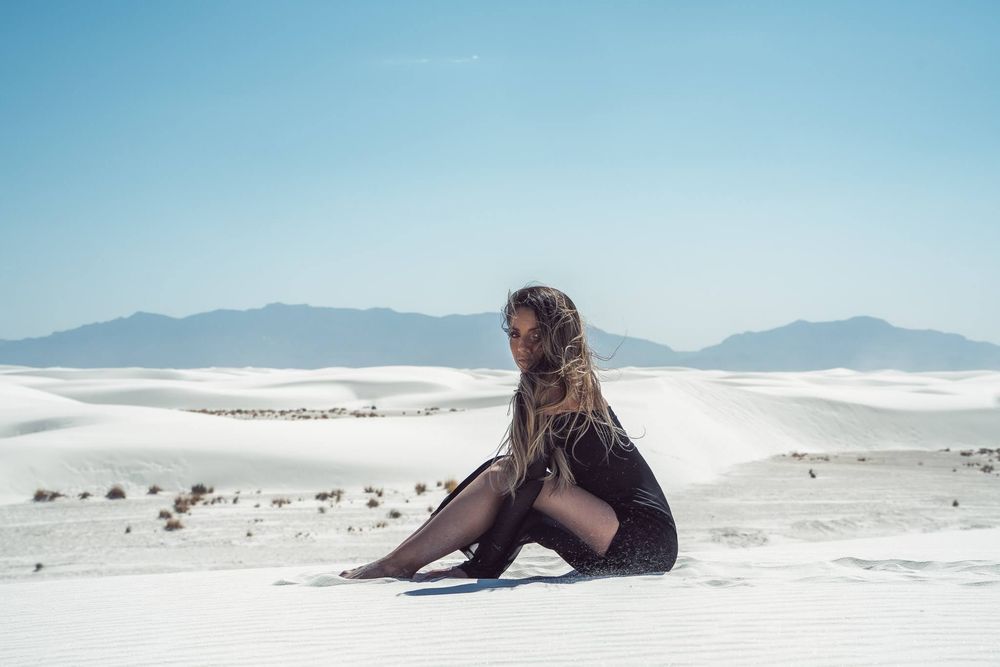 White sand dunes stretch across a desert landscape with distant mountains under a bright blue sky.