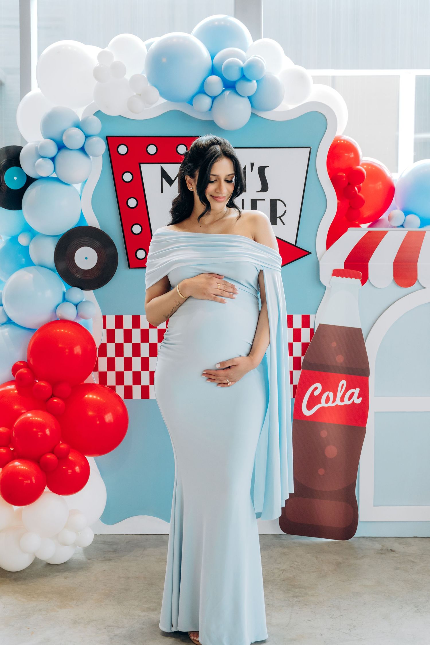 A maternity photoshoot with retro diner decor featuring red and blue balloon garlands and vintage Coca-Cola themed backdrops.