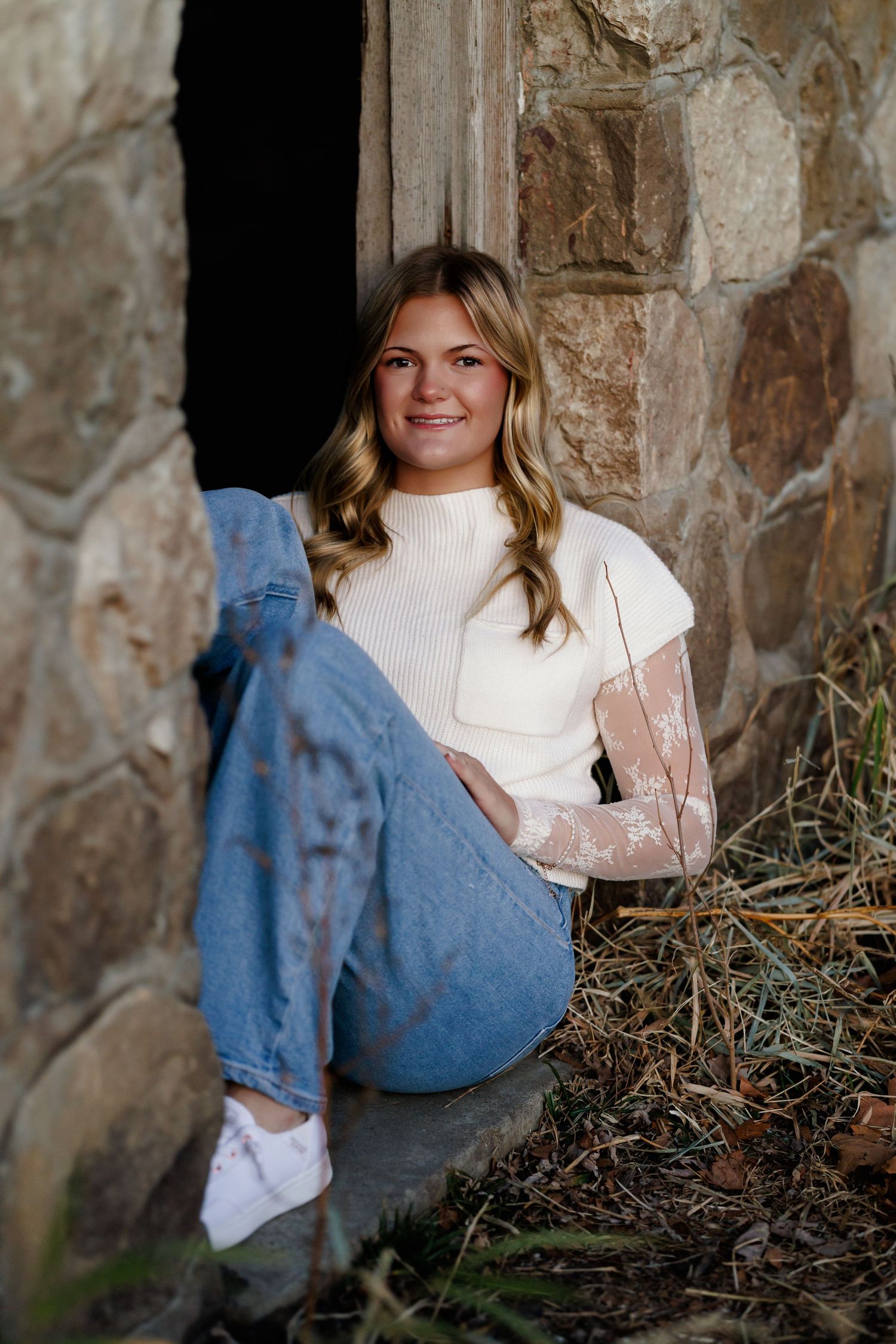 A senior girl in a white top sits against a rustic stone wall while casting a blue shadow in natural light.