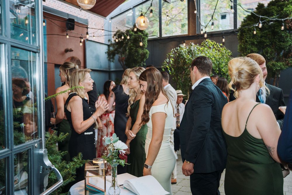 Guests mingle and celebrate at an elegant indoor wedding reception surrounded by lush greenery and warm string lights.