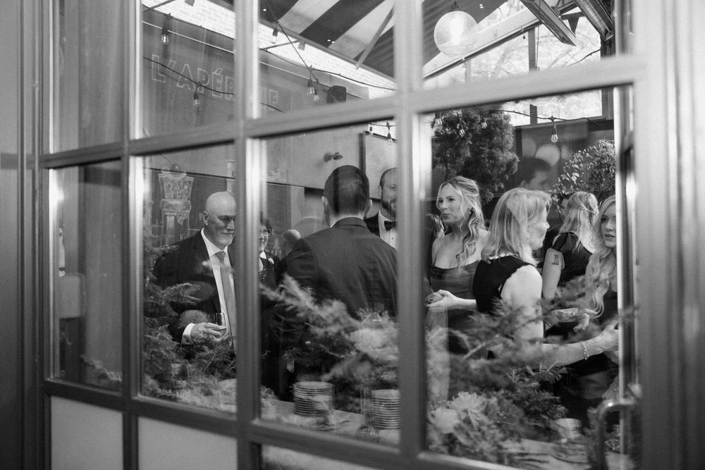 Black and white photo of wedding guests socializing in a greenhouse venue, viewed through glass window panes.