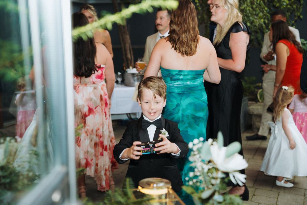 Young boy in a tuxedo holds a camera at an elegant outdoor wedding reception with guests mingling.