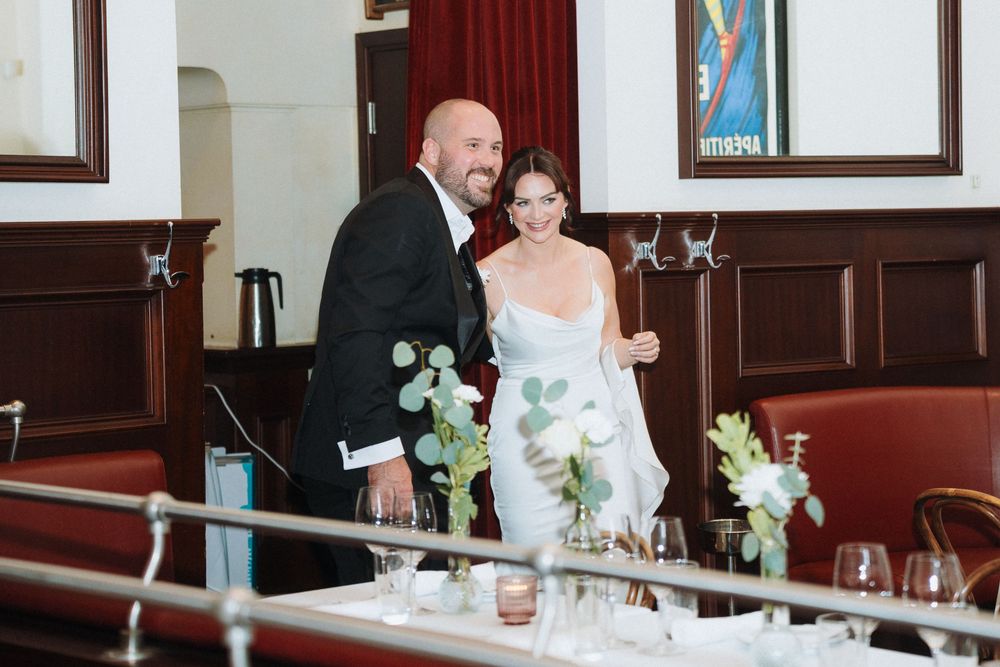 Smiling bride in white gown and groom in black tuxedo standing together at their wedding reception venue.