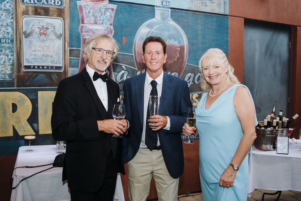 Three guests pose smiling with wine glasses in front of a colorful mural at a wedding.