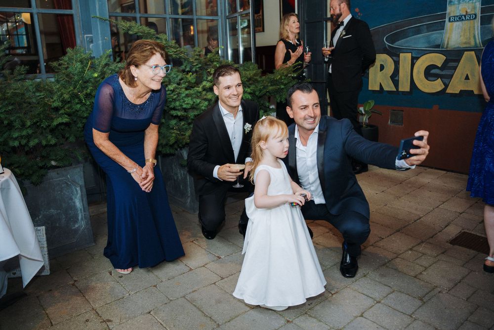 Wedding guests take selfie with adorable flower girl in white dress at outdoor reception venue.