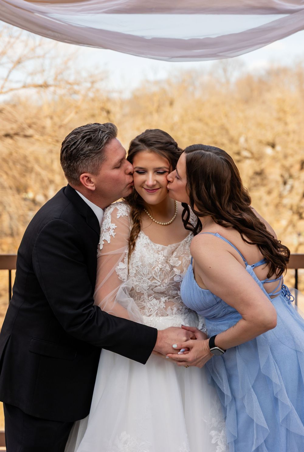 Bride in lace wedding gown receives kisses on cheeks from parents in black suit and blue dress at outdoor ceremony.