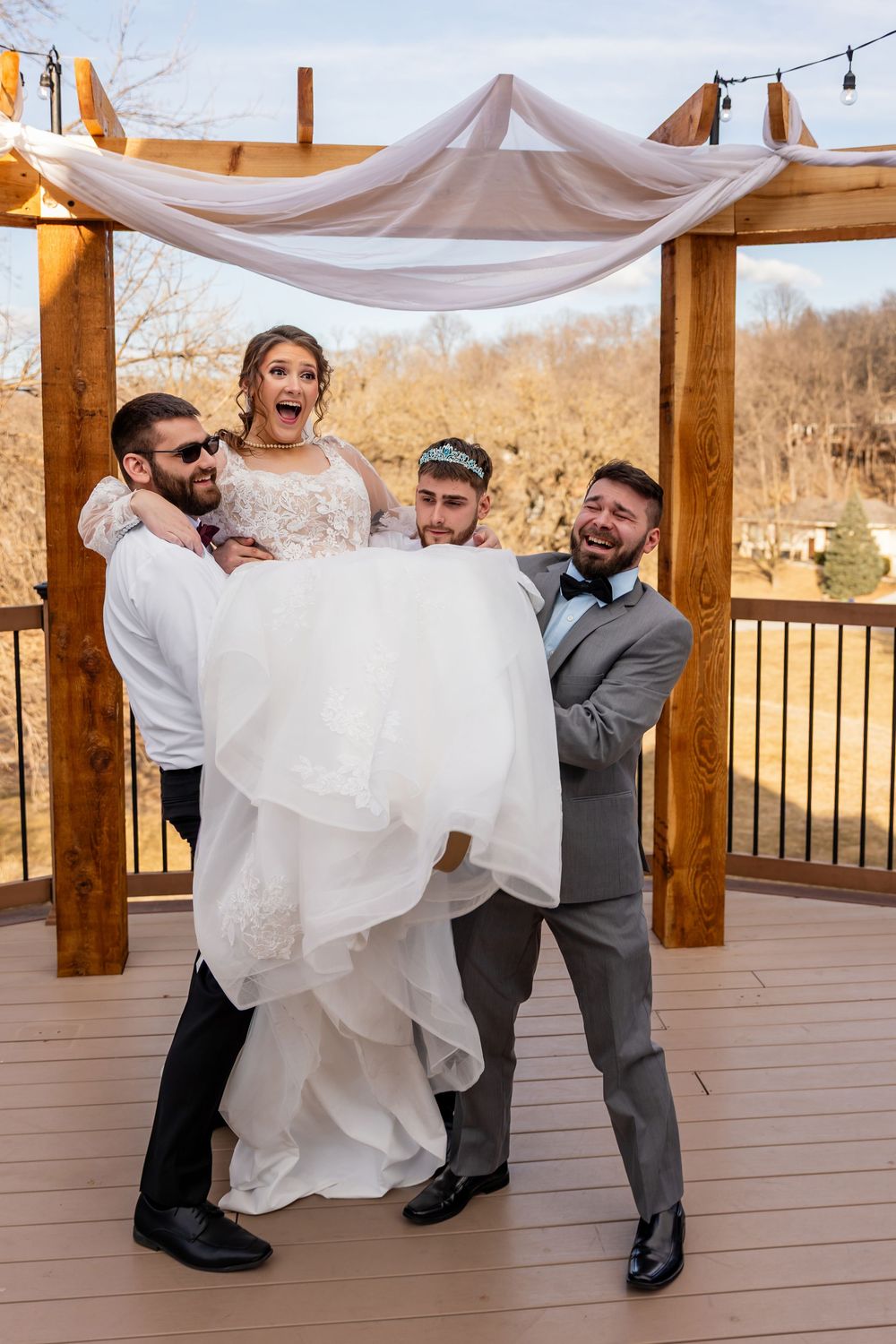 Bride in white gown lifted by groomsmen under decorated pergola, laughing joyfully at outdoor wedding celebration.