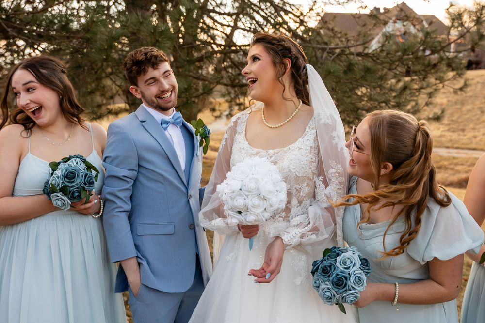 Bride in white lace gown laughing with groom in blue suit and bridesmaids in blue dresses holding floral bouquets outdoors.