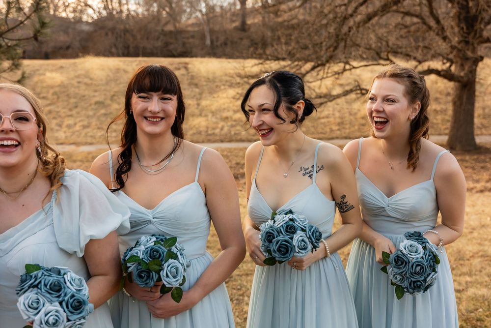 Four bridesmaids in light blue dresses laughing and holding blue rose bouquets outdoors at a wedding.