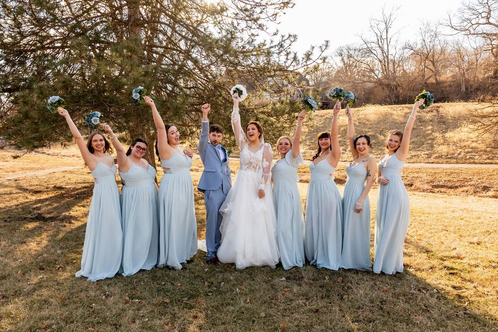 Bride and groom celebrate with seven bridesmaids in light blue gowns, raising bouquets outdoors at a wedding.