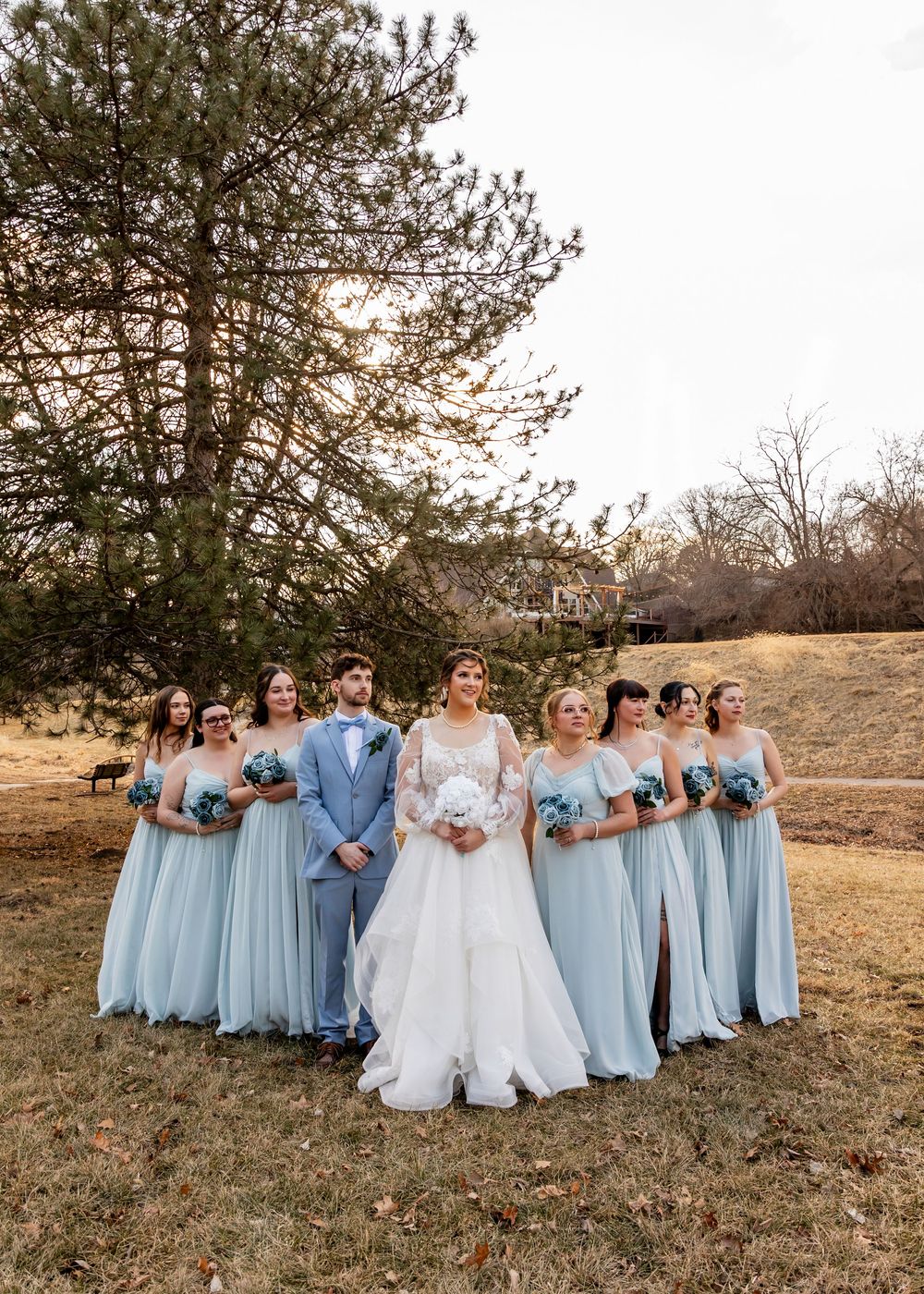 Bride and groom with bridesmaids in light blue gowns posing outdoors near pine trees on a winter wedding day.