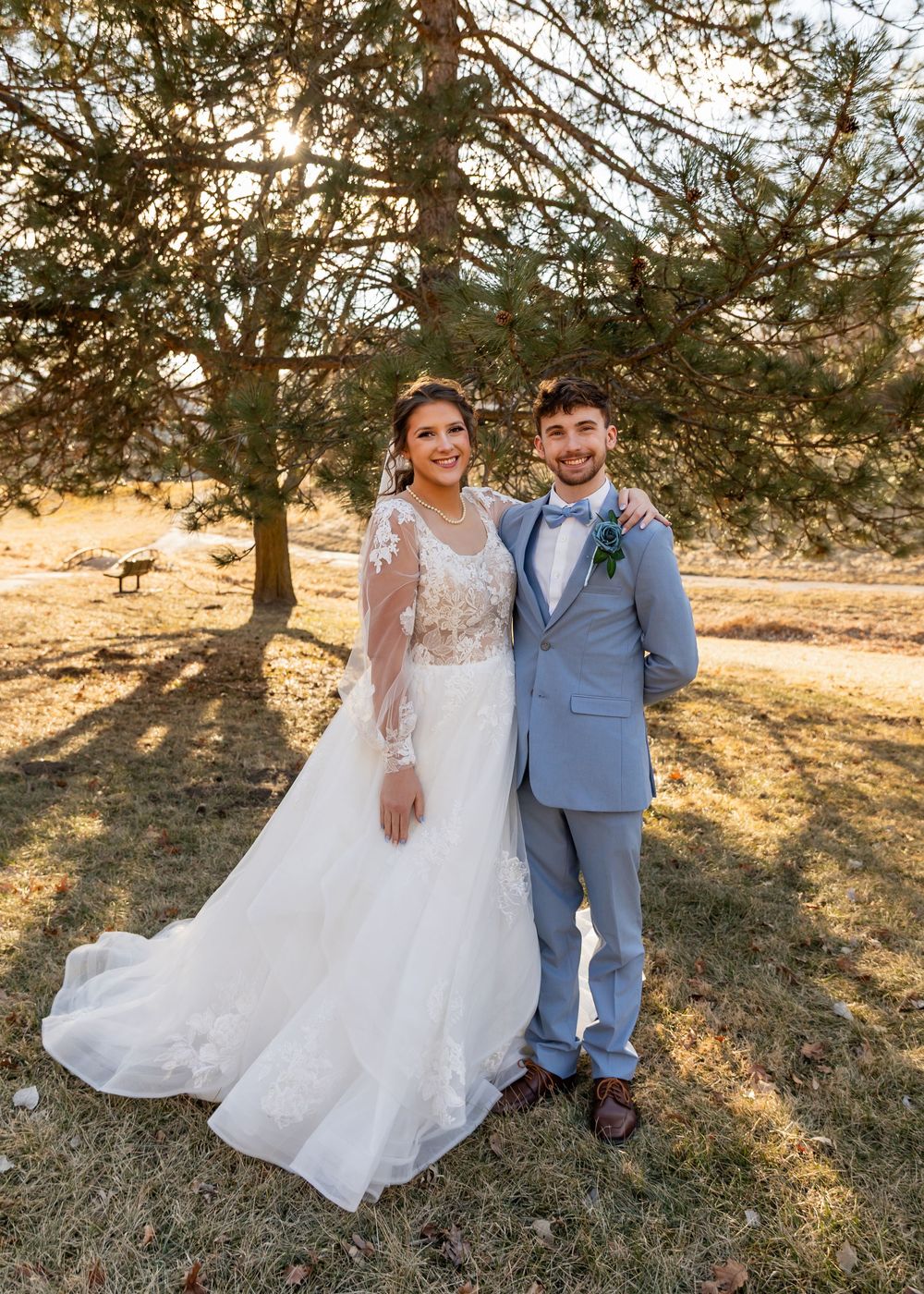 Bride in long-sleeve lace wedding gown and groom in gray suit pose together under pine trees at golden hour.