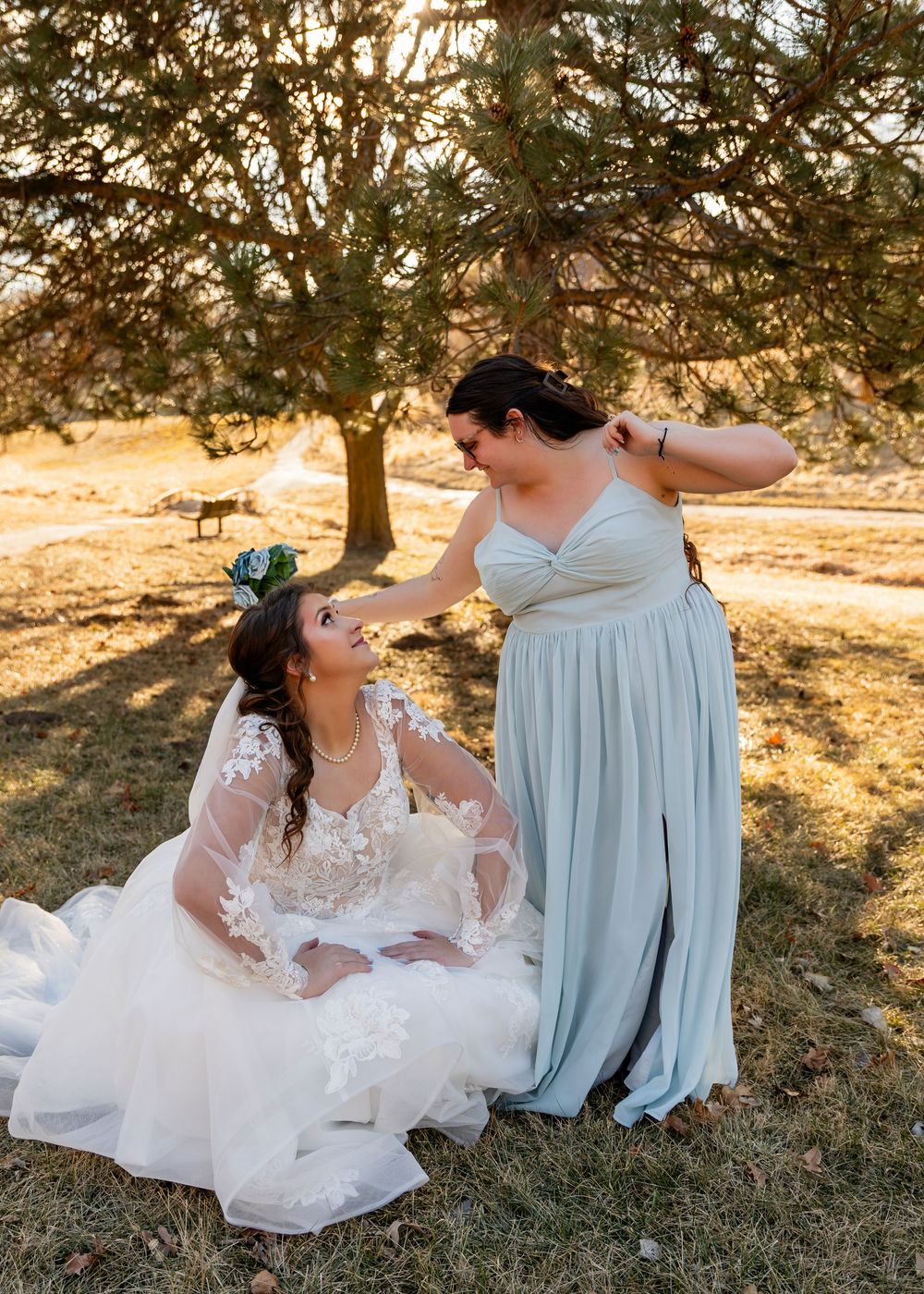 Bride in lace gown sits on grass as bridesmaid in sage green dress adjusts her hair under pine trees.