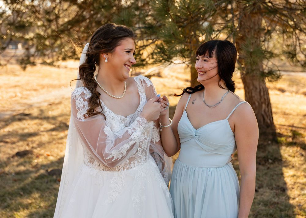 Bride in white lace gown laughs with bridesmaid in light blue dress outdoors at golden hour wedding.