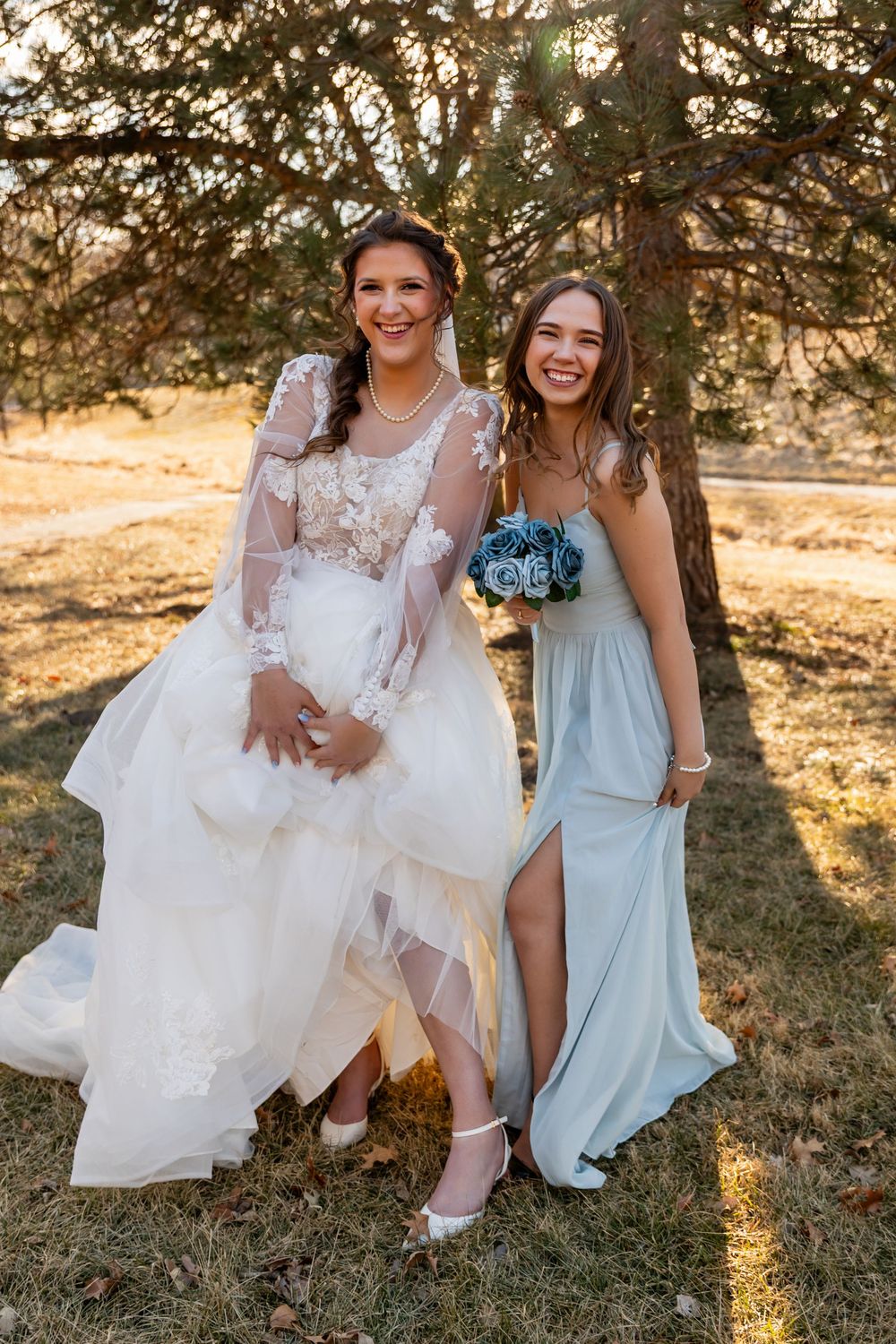 Bride in lace gown and bridesmaid in sage dress laugh together outdoors holding blue flower bouquet.