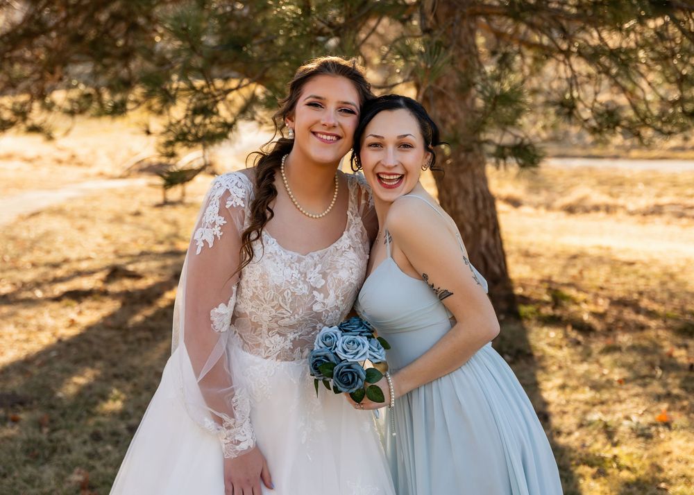 Bride and bridesmaid smiling outdoors in elegant gowns, holding blue bouquet near pine trees.