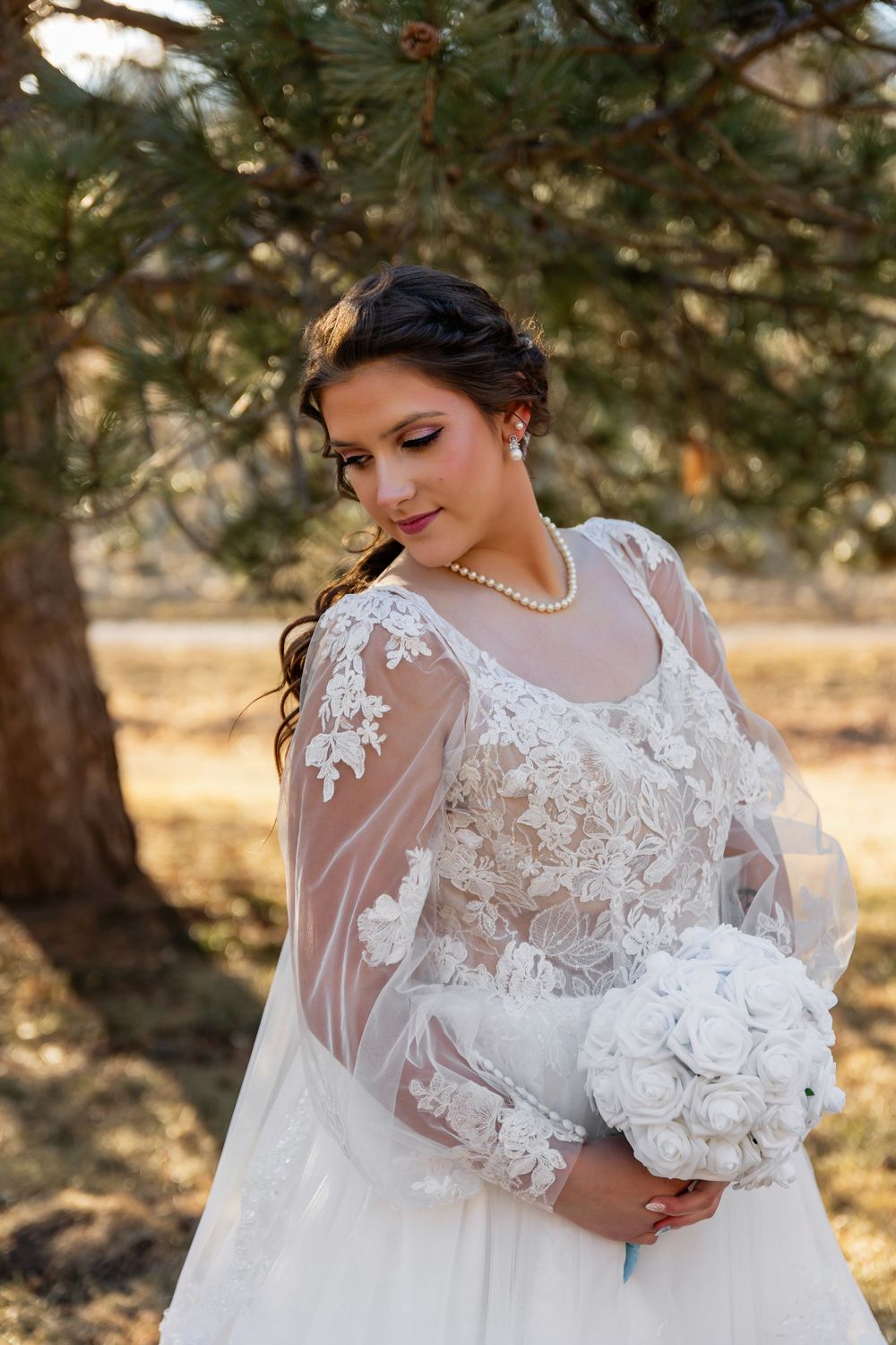 Bride in lace wedding gown with sheer sleeves holding white bouquet outdoors near pine tree.
