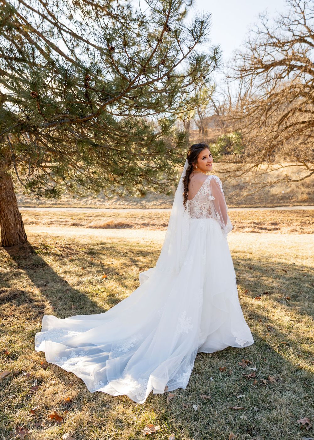 Bride in a flowing white lace wedding gown with a long train and veil, posing outdoors near pine trees.