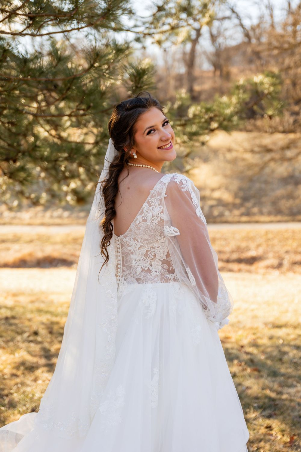 Bride smiling over shoulder showing lace back wedding dress with veil near pine tree outdoors.