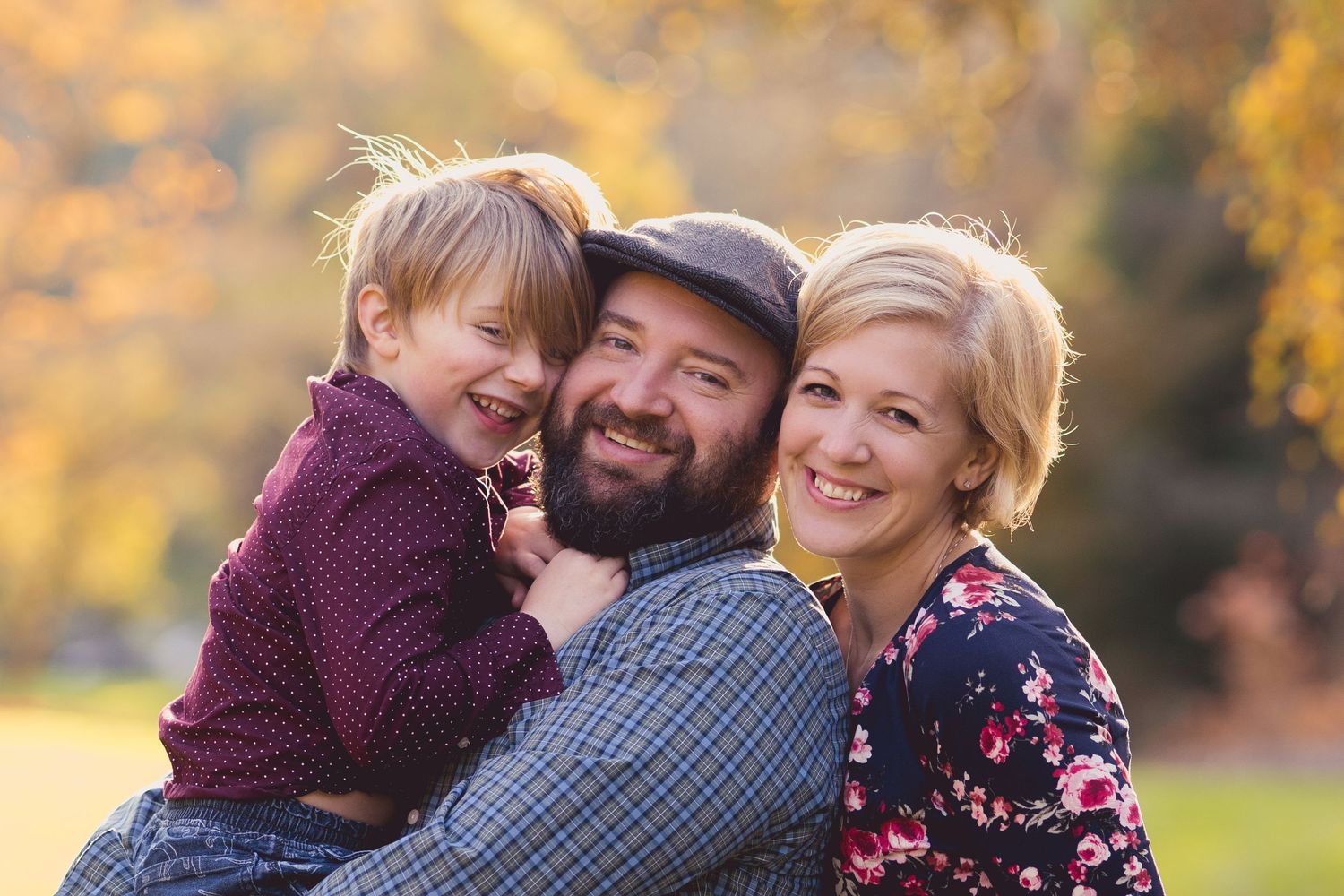 Family of three smiling outdoors in a wooded garden, parents embracing young boy in plaid shirt.