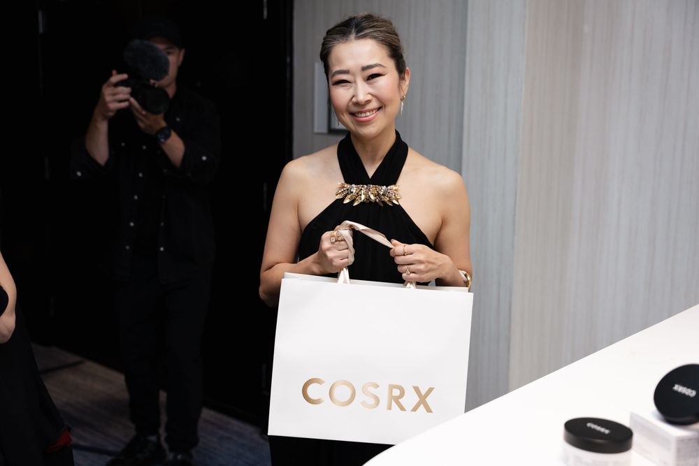 Smiling woman holds white COSRX shopping bag at elegant brand event.