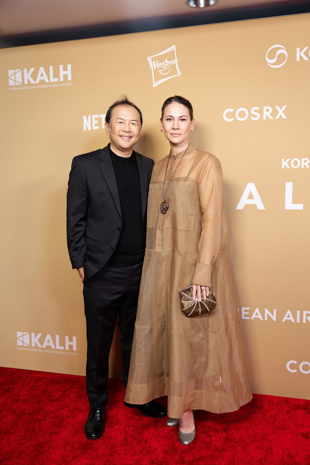 Couple poses on red carpet at Korean event wearing formal black suit and sheer gold dress.